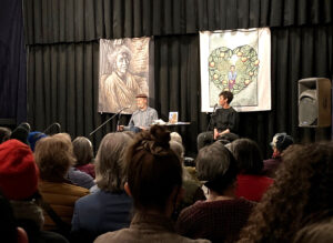 Ricardo and Autumn Brown on stage in front of a black curtain and tapestries of Ricardo's art, speaking before a large audience, as shown from the back of people's heads.